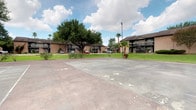 This single-story residence is part of a multi-building complex set amid mature trees, manicured lawns, and palm plantings that lend the grounds a relaxed, park-like atmosphere. The exterior common areas include a large paved recreational court—suitable for tennis or basketball—illuminated by a tall floodlight pole, with the surrounding brick buildings providing a cohesive architectural backdrop.

Inside, the interior reflects a thoughtfully curated aesthetic. The dining area serves as a focal point of the space, anchored by a striking round green marble table paired with rattan chairs atop a woven area rug. Dark concrete-style flooring runs throughout, grounding the neutral, cream-toned walls and lending an understated industrial quality to the overall design. Recessed lighting provides consistent overhead illumination.

A distinctive sliding barn door separates the dining area from an adjoining corridor or room, adding architectural character and functional flexibility. Framed artwork displayed on the walls—including a large abstract piece in muted tones—reinforces the gallery-like sensibility of the interior. Glimpses through doorways reveal additional rooms decorated with framed prints, hinting at a cohesive, design-forward approach throughout the residence.