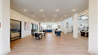 Belltower Lounge at 3400 Tevis Dr. in Louisville, KY is a generously sized, two-story community amenity space designed to offer residents a versatile and modern environment for relaxation, socializing, and recreation. The ground floor welcomes visitors through a staffed reception area with a warm wood-toned desk, leading into expansive open lounge zones finished with light hardwood-style flooring and crisp white walls. Seating areas are thoughtfully arranged with contemporary sofas and accent chairs in navy, coral, and earth tones. Game amenities include a shuffleboard table and a pool table, while a stone-surround fireplace with a mounted TV adds warmth to one lounge corner. A fully equipped kitchen features white shaker cabinetry, granite countertops, subway tile backsplash, and stainless steel appliances. Abundant natural light streams through large windows and glass-paneled doors that open to an outdoor terrace. The second floor, visible from below through dramatic floor-to-ceiling steel-framed glass panels, houses a well-appointed fitness center with a full suite of cardio equipment. A sleek staircase with a metal handrail connects both levels, and curated artwork throughout adds a refined, community-focused character to the entire space.