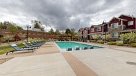 This outdoor pool area is a well-appointed amenity space designed for relaxation and recreation. The centerpiece is a large rectangular swimming pool with clear blue water, surrounded by an expansive concrete deck that provides ample room for circulation and leisure. A row of blue lounge chairs lines the poolside, offering dedicated spaces for sunbathing and unwinding.

The area is fully enclosed by a black wrought iron fence, providing both security and a defined boundary for the space. Access is controlled through a gated entry, which displays safety signage including a life ring station mounted nearby. The surrounding buildings feature a distinctive craftsman-inspired aesthetic, combining rich red board-and-batten siding with stacked stone masonry accents, contributing a warm, residential character to the outdoor setting.

A pergola structure on one end of the pool deck provides a shaded gathering spot, complemented by additional seating. On the opposite side, a designated grilling area with stainless steel grills and manicured lawn patches enhances the space's functionality for outdoor entertaining. Landscaping throughout includes ornamental trees, flowering shrubs, and ground cover plantings that soften the hardscape and add seasonal color to the overall environment.