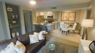 This single-floor residence showcases a thoughtfully designed open-concept layout that unifies the living, dining, and kitchen areas into one cohesive and functional space. The living area is anchored by a plush dark sofa accented with decorative throw pillows, a glass-top coffee table, and a patterned area rug, creating a warm and inviting atmosphere. A ceiling fan with an integrated light fixture provides both comfort and ambient illumination overhead.

The kitchen is well-equipped with stainless steel appliances, including a refrigerator, microwave, and range, all complemented by a classic mosaic tile backsplash and light-toned countertops. Gray cabinetry adds a contemporary feel, while a raised breakfast bar with bar seating offers a casual dining option and serves as a natural divider between the kitchen and living space.

Adjacent to the kitchen, the dining area accommodates a full table and chairs set beneath a piece of large-format wall art, adding a touch of elegance to the space. The neutral color palette of warm grays and beiges flows consistently throughout, lending the home a modern yet comfortable ambiance. A hallway visible from the main area suggests additional private spaces beyond the open living zone.