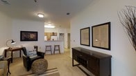 This single-floor residence offers a well-designed open-concept layout that seamlessly connects the living and kitchen areas, creating a comfortable and functional living environment. The living space features neutral-toned carpeting that adds warmth underfoot, while crown molding along the ceiling provides a refined architectural touch throughout. Decorative framed artwork adorns the white walls, complementing a dark wood console that anchors the room with a sense of elegance. The seating area is thoughtfully arranged with a leather accent chair and woven ottoman, contributing to an inviting, casual ambiance. A peninsula breakfast bar separates the kitchen from the living space, lined with wooden bar stools that encourage informal dining and social interaction. The kitchen is equipped with classic wood-toned cabinetry, a white refrigerator, and ample counter space, offering a practical and efficient cooking environment. Flush-mount ceiling light fixtures illuminate both the living and kitchen areas evenly. A hallway visible beyond the kitchen hints at additional private spaces within the unit. The overall design balances functionality and comfort, making this a practical and welcoming single-floor home.