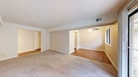 This single-floor space showcases an open and functional layout designed to maximize usable living area. The main living space features neutral-toned carpet flooring that transitions to hardwood flooring in an adjoining section, creating a natural visual distinction between zones. Warm beige walls complement the light-colored textured ceiling throughout, lending a cohesive and understated aesthetic to the interior.

A ceiling fan with an integrated light fixture is installed in the area with hardwood flooring, offering both air circulation and ambient lighting. A window positioned along one wall allows natural light to filter into the space, brightening the hardwood-floored section. HVAC venting is visible in the ceiling, indicating a central air system.

The layout opens toward multiple adjoining areas, including what appears to be a kitchen — identifiable by a white refrigerator visible through a doorway — as well as additional rooms accessible through separate openings on the opposite side. White trim and baseboards run consistently throughout, adding a clean, finished look. Overall, the space presents a straightforward, practical interior well-suited for comfortable everyday living.