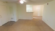 This single-floor space features an open-concept layout that connects the main living area to a secondary space, creating a sense of continuous flow throughout. The living area is finished with wall-to-wall neutral beige carpeting, which extends into the adjoining room, lending a cohesive and understated aesthetic throughout the unit.

A ceiling fan with a light fixture is mounted centrally in the main living area, providing both air circulation and ambient lighting. A closet door is visible to one side, offering practical storage within easy reach of the main living space.

The kitchen is positioned centrally and features a raised pass-through counter that opens to the living area, making it functional for casual dining or entertaining. White cabinetry and a built-in microwave are visible within the kitchen, contributing to a clean, uniform look. The kitchen is illuminated with overhead lighting.

The adjoining room, separated by a partial wall, is accented by an elegant chandelier, adding a decorative touch. The walls throughout are finished in a soft, neutral tone, complementing the light carpeting and creating a bright, welcoming atmosphere across the entire space.