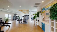 Smith Creek Health Hub is a well-appointed, single-story fitness facility located at 4645 Gordon Rd in Wilmington, NC. The space is designed to accommodate a full spectrum of fitness activities, with distinct zones thoughtfully arranged across an open, flowing floor plan. Warm hardwood-style flooring runs throughout, creating a cohesive and inviting atmosphere.

The main gym area is the centerpiece of the facility, featuring a striking coffered ceiling with warm wood inlays, ceiling fans, and oversized garage-style windows that flood the space with natural light and offer outdoor views. This area houses a comprehensive selection of Precor strength machines, cable systems, and cardio equipment, arranged to allow ample room for movement between stations.

A dedicated free weights zone offers a full dumbbell rack, a power rack with weight plates, and a motivational typography mural on the wall, reinforcing the facility's wellness-focused identity. Adjacent to the main floor, a bright cycling and group fitness studio features Spinner bikes, stability balls, wall-mounted displays, and direct access to an outdoor area through glass doors.

A separate yoga and studio room provides a serene, open space finished in soft sage green tones, with ceiling fans, mirrors, modular shelving, and natural light from high windows — ideal for yoga, stretching, or small group classes. Together, these spaces create a comprehensive, welcoming fitness environment.