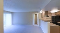 This single-floor residential unit offers a straightforward and functional layout designed for comfortable everyday living. The main living area is finished with wall-to-wall carpeting in a neutral tone, creating a soft and cohesive foundation throughout the space. Walls are painted in a warm beige, complementing the light-colored carpet and contributing to a clean, understated aesthetic. The textured ceiling adds a subtle layer of detail overhead, while a ceiling-mounted light fixture provides overhead illumination.

A standout feature of the living area is a built-in closet with a hanging rod, offering dedicated storage directly accessible from the main room. The open doorway leading to this closet enhances the sense of connectivity within the space.

To one side, a kitchen area is partially visible, separated from the living space by a raised counter that functions as a breakfast bar or pass-through. Overhead cabinetry in a warm wood tone provides ample kitchen storage, while the countertop surface appears to feature a stone or laminate finish. A ceiling-mounted light above the kitchen area ensures the workspace is well-lit.

Ventilation is provided via a ceiling vent, and multiple electrical outlets are positioned along the walls for practical use. Overall, the unit presents a move-in-ready, no-frills living environment with efficient use of space.