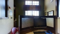 This single-floor office space is designed with productivity and focus in mind. The room is distinguished by its deep, dark green walls, which create a grounded and professional atmosphere. Natural light filters in through two stacked windows fitted with blinds, offering brightness while maintaining privacy and light control throughout the day.

The workspace is anchored by a substantial dark wood desk with a built-in privacy panel, providing a sense of separation and concentration within the room. The desk is equipped with a desktop computer and keyboard, making it a fully functional workstation. To the left, built-in cabinetry and shelving offer ample storage for books, supplies, and personal items, contributing to an organized and tidy environment.

A pendant lamp with a patterned shade hangs above, adding a stylish decorative touch that complements the room's color palette. The flooring appears to be a light-toned tile, offering a pleasant contrast to the darker wall tones. A seating area is partially visible, suggesting the space accommodates both individual work and brief consultations. Overall, this office combines practical design with a warm, focused ambiance suited for professional use.