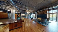 Located on Charlton Street in Everett, Massachusetts, this single-floor lounge showcases a compelling blend of industrial and warm, organic design elements. Polished hardwood floors stretch across the expansive open layout, contrasting beautifully with the raw exposed concrete ceilings, structural columns, and visible ductwork overhead — hallmarks of thoughtful industrial-chic architecture.

The space is anchored by a striking fireplace feature wall, dramatically stacked floor-to-ceiling with split logs alongside a sleek linear hearth, creating a bold focal point. Adjacent to it, a seating area with plush armchairs and a shag area rug invites relaxed gathering, while large multi-pane windows bathe the zone in natural light and offer views of the surrounding greenery.

A secondary lounge nook is defined by a rich cobalt blue rug and furnished with leather sofas and accent chairs, complemented by reclaimed wood wall paneling and curated textile artwork. A dedicated refreshment station, outfitted with natural wood cabinetry and a built-in Starbucks coffee machine, provides convenience for occupants.

Near the entrance, a substantial dark wood reception counter serves as a welcoming focal point, framed by an open transitional area with polished concrete flooring. The overall ambiance is warm, sophisticated, and purposefully eclectic — an inviting communal space designed for comfort and community.