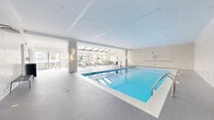 Located at 1254 Hennepin Ave N in Minneapolis, this generously sized indoor aquatic and lounge facility offers a refined, resort-style experience in the heart of the city. The space is anchored by a large, rectangular lap pool finished in crisp white tile, its vivid blue water reflecting the abundant natural light that streams in from multiple large windows and a dramatic skylight structure overhead. The pool deck is clad in fine white mosaic tile throughout, lending the entire space a clean, cohesive aesthetic.

Adjacent to the pool, a dedicated sunning and lounge area sits beneath a striking skylight roof, filled with cushioned chaise lounges and cafe-style seating. Floor-to-ceiling windows in this area frame panoramic views of the Minneapolis downtown skyline, creating a bright, airy retreat regardless of the season. Warm gold wall sconces accent the neutral stone-tiled walls, adding a touch of elegance to the contemporary design.

The facility also features a separate pool entry area finished in textured tile, with glazed doors providing connectivity to adjoining amenity spaces. The overall ambiance is modern and sophisticated, balancing functionality with a polished, upscale aesthetic suited for both lap swimming and leisurely relaxation.