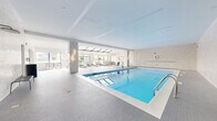 Located at 1254 Hennepin Ave N in Minneapolis, this generously sized indoor aquatic and lounge facility offers a refined, resort-style experience in the heart of the city. The space is anchored by a large, rectangular lap pool finished in crisp white tile, its vivid blue water reflecting the abundant natural light that streams in from multiple large windows and a dramatic skylight structure overhead. The pool deck is clad in fine white mosaic tile throughout, lending the entire space a clean, cohesive aesthetic.

Adjacent to the pool, a dedicated sunning and lounge area sits beneath a striking skylight roof, filled with cushioned chaise lounges and cafe-style seating. Floor-to-ceiling windows in this area frame panoramic views of the Minneapolis downtown skyline, creating a bright, airy retreat regardless of the season. Warm gold wall sconces accent the neutral stone-tiled walls, adding a touch of elegance to the contemporary design.

The facility also features a separate pool entry area finished in textured tile, with glazed doors providing connectivity to adjoining amenity spaces. The overall ambiance is modern and sophisticated, balancing functionality with a polished, upscale aesthetic suited for both lap swimming and leisurely relaxation.