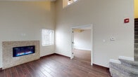 This two-floor residence offers a thoughtfully designed layout with a range of distinctive architectural details throughout. The main living area immediately impresses with its soaring vaulted ceilings that amplify the sense of space and openness. Multiple windows with blinds allow natural light to fill the room, while rich, dark wood-look tile flooring runs consistently through the main level, adding warmth and visual continuity. A standout feature of the living area is the built-in electric fireplace set within a stone tile surround, providing both a functional focal point and an elegant design element. The galley-style kitchen is outfitted with rich dark cabinetry featuring frosted glass accents, white countertops, a stainless steel sink, stainless steel appliances, and a horizontally striped tile backsplash that adds texture and depth. The main level also includes a full bathroom featuring a wood-toned vanity with an integrated white sink, a backlit mirror, and a shower-tub combination. The upper floor offers a carpeted room with a charming sloped ceiling that follows the roofline, along with a window that brings in natural light. A second full bathroom on this level mirrors the clean, modern aesthetic found below. Throughout, the neutral warm-toned walls and clean white trim create a cohesive and inviting atmosphere.