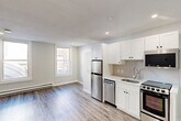 This 290 square foot studio apartment, located at 76 Franklin St in Worcester, MA, offers a thoughtfully designed living space on a single floor. The interior showcases a modern aesthetic with a cohesive color palette of whites and neutrals. The living area is open and spacious, featuring wood-look vinyl flooring that extends throughout the apartment, creating a sense of continuity and warmth.

The kitchen is a standout feature, equipped with sleek stainless steel appliances including a refrigerator, dishwasher, and range. White Shaker-style cabinetry provides ample storage and contributes to the bright, clean look of the space. A window above the kitchen sink allows natural light to filter in, enhancing the room's airy feel.

The living room area is versatile, with enough space to accommodate both sleeping and seating arrangements. Large windows flood the room with natural light and offer views of the surrounding area. Recessed lighting in the ceiling provides additional illumination options.

The bathroom, while compact, is efficiently designed with a modern white vanity, toilet, and what appears to be a shower stall (not fully visible in the image). The continuation of the wood-look flooring into the bathroom maintains the cohesive design throughout the apartment.

Overall, this studio apartment maximizes its 290 square feet through smart design choices, creating a functional and aesthetically pleasing living space ideal for a single occupant or couple.