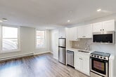 This 290 square foot studio apartment, located at 76 Franklin St in Worcester, MA, offers a thoughtfully designed living space on a single floor. The interior showcases a modern aesthetic with a cohesive color palette of whites and neutrals. The living area is open and spacious, featuring wood-look vinyl flooring that extends throughout the apartment, creating a sense of continuity and warmth.

The kitchen is a standout feature, equipped with sleek stainless steel appliances including a refrigerator, dishwasher, and range. White Shaker-style cabinetry provides ample storage and contributes to the bright, clean look of the space. A window above the kitchen sink allows natural light to filter in, enhancing the room's airy feel.

The living room area is versatile, with enough space to accommodate both sleeping and seating arrangements. Large windows flood the room with natural light and offer views of the surrounding area. Recessed lighting in the ceiling provides additional illumination options.

The bathroom, while compact, is efficiently designed with a modern white vanity, toilet, and what appears to be a shower stall (not fully visible in the image). The continuation of the wood-look flooring into the bathroom maintains the cohesive design throughout the apartment.

Overall, this studio apartment maximizes its 290 square feet through smart design choices, creating a functional and aesthetically pleasing living space ideal for a single occupant or couple.