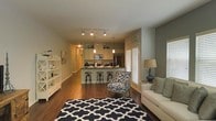 This single-floor residence showcases an open-concept layout that integrates the kitchen, dining, and living areas into one cohesive, flowing space. The kitchen is appointed with cream-toned cabinetry, stainless steel appliances including a microwave and refrigerator, and a dark-topped breakfast bar with industrial-style metal stools, creating a functional yet stylish cooking and casual dining zone. Pendant lighting drops elegantly above the bar area, complementing the track lighting overhead that illuminates the space evenly. The living area is anchored by a bold navy and white geometric-patterned area rug, adding a striking visual contrast against warm hardwood flooring that extends throughout. A neutral sofa with accent pillows and a patterned accent chair provide comfortable seating, while a floor lamp with a warm-toned shade adds ambiance. White plantation shutters on multiple windows allow for controlled natural light. An open shelving unit along one wall offers display and storage space. The overall ambiance is warm and inviting, combining contemporary design elements with practical furnishings.