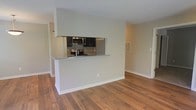 This single-floor residence offers a clean, open-concept layout designed for comfortable everyday living. The space is anchored by warm hardwood flooring that flows seamlessly throughout, lending a cohesive and inviting feel to the interior.

The kitchen is positioned behind a half-wall pass-through that features a granite countertop, creating a natural divide between the cooking area and the open living space beyond. Dark cabinetry provides a striking contrast against the light-toned walls, while stainless steel appliances — including a microwave and electric range — offer a modern, functional aesthetic. The pass-through design allows for easy interaction between the kitchen and the adjoining living area, making the layout well-suited for both everyday use and casual entertaining.

The living area is bright and open, with neutral, soft gray-green walls that contribute to a calm and airy atmosphere. White baseboards add a crisp finishing touch throughout. A louvered door, likely leading to a utility or storage closet, can be spotted near the kitchen, adding practical storage to the space. Ventilation registers are neatly placed along the walls, reflecting a thoughtfully maintained interior. Overall, the space presents a simple yet refined aesthetic with a welcoming, move-in-ready character.