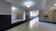 This single-floor space presents a clean, open interior defined by a series of striking design elements. The most prominent feature is a full wall of floor-to-ceiling mirrors along one side, which visually expands the room and reflects natural light throughout the space, creating a bright and airy atmosphere. The polished terrazzo flooring adds a classic, durable finish underfoot with a subtle speckled pattern that contributes to the space's refined character.

Running parallel to the mirrored wall is an elongated windowed corridor, lined with large windows that allow generous amounts of natural light to filter in while offering views of an adjacent outdoor balcony or walkway. This arrangement gives the space a sense of considerable depth and length. Track lighting installed along the corridor ceiling supplements the natural light effectively.

Overhead, an elegant crystal flush-mount light fixture serves as a decorative focal point, lending a touch of sophistication to the otherwise understated interior. The neutral gray walls and white ceiling maintain a clean, versatile backdrop. The overall ambiance is polished and functional, making the space well-suited for a variety of uses, from fitness or dance studios to event or exhibition venues.