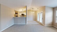 This thoughtfully designed single-floor residence offers a comfortable and functional layout across a well-utilized footprint. The open-concept living and kitchen area is the heart of the home, featuring vaulted ceilings that enhance the sense of volume and space. Pendant chandeliers illuminate both the kitchen and living zones, adding a warm, refined touch. The kitchen is equipped with stainless steel appliances, including a refrigerator and range, complemented by crisp white shaker cabinetry, light countertops, and durable wood-look flooring. A raised breakfast bar separates the kitchen from the carpeted living area, which is anchored by a tile-surround fireplace and framed by large windows and glass French doors that invite abundant natural light and outdoor views. The spacious bedroom features vaulted ceilings, soft carpet, a ceiling fan, a generous window overlooking the outdoors, and access to a dedicated closet with additional storage. A hallway connects the main living areas to the bedroom and full bathroom, which includes a tiled tub-shower combination with chrome fixtures. Neutral tones and clean finishes are carried throughout, creating a cohesive and inviting atmosphere well-suited for comfortable everyday living.