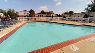 This community amenity space offers a comprehensive recreational experience both outdoors and indoors. At its centerpiece is a generously sized outdoor swimming pool with clear turquoise water, framed by classic brick coping and a spacious concrete sun deck lined with blue lounge chairs — ideal for relaxing under open skies. The pool area is enclosed by a white vinyl fence, providing privacy while maintaining views of surrounding mature trees. Adjacent to the pool, a tennis court adds an additional layer of outdoor recreation for residents.

The indoor clubhouse complements the outdoor amenities with a warm and welcoming interior. The lounge area features hardwood-style flooring in a herringbone pattern, a fireplace with a wall-mounted television, and comfortable seating arranged for social gatherings. A teal accent wall adds a pop of color and visual interest throughout the space. The dining area includes vaulted ceilings, framed wall art, and a water cooler station for convenience. A fully equipped kitchen with warm wood cabinetry, stainless steel appliances, a breakfast bar, and ample counter space rounds out the interior. Large windows allow natural light to flow freely throughout the clubhouse, creating a bright and inviting atmosphere.