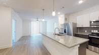 This single-floor residence showcases a thoughtfully designed open-concept layout that seamlessly connects the kitchen and living areas. The kitchen is a standout feature, equipped with stainless steel appliances including a French door refrigerator with ice maker, electric range, and built-in microwave. A generous granite-topped island with an undermount sink and pendant lighting serves as the centerpiece, complemented by two-tone cabinetry featuring white upper cabinets and dark espresso lower cabinets, accented by a textured tile backsplash.

Light wood-look flooring runs throughout the space, enhancing the bright, airy atmosphere created by crisp white walls and recessed lighting. The living area benefits from a ceiling fan and access to natural light through vertical blinds. Built-in shelving near the kitchen provides additional storage and display options.

The bathrooms offer both practical and comfortable features. One bathroom includes a tub-shower combination with full-height tile surround and a natural light window, while another features a glass-enclosed shower with accessibility grab bars. Both bathrooms maintain the light, clean aesthetic found throughout the rest of the space, with wood-look flooring continuing seamlessly from the main living areas.