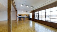 This loft offers a generously sized, open-concept single-floor layout that seamlessly blends industrial character with modern functionality. The space is defined by its exposed brick accent walls, which add warmth and texture alongside polished hardwood floors that reflect natural light throughout the interior. Large, multi-pane industrial windows line one wall, drawing in abundant daylight and providing views of the surrounding area, while contributing to the loft's authentic urban aesthetic.

The kitchen is positioned along one wall and features warm wood cabinetry, stainless steel appliances including a range, microwave, and refrigerator, and ample counter space. Track lighting and a multi-arm pendant fixture provide versatile overhead illumination throughout the open living area, complementing the natural light from the oversized windows.

Structural columns punctuate the space, reinforcing the loft's industrial heritage while helping to define zones within the open floor plan. The high ceilings enhance the sense of volume and airiness, making the space feel expansive and flexible. Overall, this loft presents a distinctive combination of raw architectural detail and comfortable, livable design.