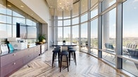 Located in the heart of Tysons at 1755 Tysons Central Street, this stunning amenity lounge delivers an exceptional blend of modern design and refined comfort. The single-floor space is defined by dramatic floor-to-ceiling curved glass curtain walls that flood the interior with abundant natural light and frame sweeping panoramic views of the Tysons skyline and surrounding greenery.

Soaring double-height ceilings amplify the sense of openness throughout, while elegant crystal and sculptural chandeliers add a touch of luxury from above. The herringbone-patterned hardwood flooring serves as a cohesive design element across the entire space, lending warmth and sophisticated texture underfoot.

The layout is thoughtfully organized into distinct social zones. A lounge area features contemporary seating arrangements with plush blue accent chairs and sleek round tables, ideal for casual gatherings. A dedicated dining area accommodates multiple seating configurations with modern tables and chairs. A wet bar and catering station with dark cabinetry and a high-top communal table with bar stools provides a stylish hub for entertaining.

A gallery-style accent wall showcases a curated collection of eclectic artwork, adding artistic character to the space. Direct access to an outdoor terrace with seating further extends the social experience, offering open-air views of the Tysons corridor.