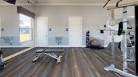 This single-floor fitness center is thoughtfully designed to support a full range of workout routines. The main exercise area is generously sized, featuring dark-toned, multi-striped carpet flooring that adds a modern aesthetic throughout. Recessed lighting and crown molding lend a polished, finished look to the space.

The cardio section is well-appointed with multiple treadmills, elliptical machines, and upright bikes, all equipped with individual screens. Large windows with neutral roman shades line the walls, flooding the room with natural light and offering views of the surrounding outdoor landscape, including palm trees and community amenities.

The strength and functional training area includes a variety of cable machines, weight benches, a stability ball, medicine ball rack, and additional resistance equipment. Floor-to-ceiling mirrors expand the sense of space and support proper form during workouts. A wall-mounted television provides entertainment.

Two dedicated restrooms are accessible directly from the fitness floor, clearly marked for convenience. A kitchenette with white shaker-style cabinetry, granite countertops, a stainless steel sink, and upper and lower storage provides a practical refreshment area. A glass door offers direct access to the outdoors.