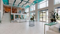 This impressive commercial space makes an immediate statement with its dramatic double-height lobby, defined by a sweeping teal-accented ceiling with undulating wave-form panels and abundant recessed lighting. Large-scale, floor-to-ceiling windows line the exterior walls, flooding the interior with natural light and offering expansive views of a palm tree-lined exterior. The polished marble-look tile flooring extends seamlessly throughout, reinforcing the clean, upscale aesthetic.

At the heart of the grand entrance foyer sits a sculptural glass-top table with an ornate metallic base, adorned with a fresh orchid arrangement that adds an elegant, welcoming touch. A cascading globe chandelier overhead serves as a luminous focal point, enhancing the sense of vertical grandeur.

The main living and lounge area features plush white sectional seating accented with ocean-blue throw pillows, conveying a sophisticated coastal ambiance. Glass-enclosed conference or office rooms are integrated directly off the lounge, offering transparency and connectivity while maintaining functional separation. A glass-railed staircase leads to a mezzanine level visible through interior glass walls, further contributing to the open, airy feel. The cohesive white-and-teal color palette, abundant natural light, and refined finishes create a polished, modern environment throughout.