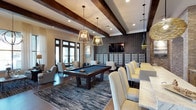 This upscale lounge is a masterfully designed social space that seamlessly blends recreation, dining, and relaxation into one cohesive environment. The interior is anchored by striking exposed dark wood ceiling beams that add warmth and architectural character, complemented by a combination of recessed lighting and statement geometric pendant fixtures that cast a welcoming ambiance throughout.

At the heart of the space sits a full-sized billiards table, positioned atop a textured area rug that defines the recreation zone. Surrounding it, plush lounge chairs offer comfortable seating for guests to gather and unwind. Large glass-paneled doors and windows line one wall, inviting abundant natural light and providing views to the exterior.

To one side, a refined dining and bar area features a long marble-topped counter flanked by tall, high-back cream upholstered chairs, ideal for social gatherings or casual dining. A tufted gray accent wall serves as an elegant backdrop to this zone, paired with a wall-mounted flat-screen television for entertainment.

The lounge also showcases a mix of exposed brick and textured dark wall panels, lending an industrial-chic aesthetic that balances sophistication with approachability. Rich dark hardwood flooring ties all the elements together, creating a stylish and inviting atmosphere suited for a variety of social occasions.