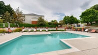 Located at 3343 Glade Creek Blvd NE in Roanoke, Virginia, this outdoor community pool area offers a well-appointed retreat designed for relaxation and recreation. The centerpiece is a generously sized swimming pool with clear turquoise water, complemented by a separate wading or spa area, making it suitable for a range of activities. The expansive pool deck is finished in smooth concrete and furnished with multiple lounge chairs and seating arrangements, including umbrella-shaded tables with orange accent chairs, providing ample space for sunbathing and socializing. The area is thoughtfully enclosed by a classic white fence accented with brick pillar posts topped with decorative terracotta planters, lending the space a charming, structured aesthetic. Lush greenery, mature trees, and manicured hedgerows surround the perimeter, offering both privacy and a pleasant natural backdrop. A small pool house or utility structure with a gabled roof is situated at one end of the deck, adding architectural character. Ornamental black iron fencing and stepped retaining walls integrate the pool area seamlessly into the broader community grounds. The overall ambiance is inviting and well-maintained, offering a comfortable outdoor amenity space with attractive landscaping and open views of the surrounding neighborhood.