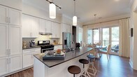 This thoughtfully designed single-floor residence showcases a cohesive blend of modern elegance and functional living. The open-concept kitchen is a standout feature, outfitted with crisp white shaker cabinetry extending to the ceiling, dark quartz countertops, subway tile backsplash, and stainless steel appliances including a gas range and French door refrigerator. A generously sized curved island with bar seating serves as the centerpiece, illuminated by cylindrical pendant lights on a track system. The adjoining dining area accommodates a round table beneath a drum pendant light and opens directly to an outdoor balcony through glass French doors, allowing abundant natural light to flow throughout. The living area offers large windows with views of lush greenery, creating a bright and inviting atmosphere. Multiple bedrooms provide comfortable retreats; one features a striking wrought-iron canopy bed frame and natural light from a large window, while a second offers a clean, neutral palette with hardwood-style flooring. The bathrooms are finished with white subway tile, a glass-enclosed shower, a soaking tub, dark quartz vanity countertops, white cabinetry, and a walk-in closet. Hardwood-style flooring flows throughout the main living areas, tying the space together with warmth and continuity.