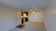 This single-floor space offers a straightforward, functional layout with an open-concept design that connects the living area directly to the kitchen. The living area is finished with wall-to-wall carpeting in a neutral beige tone, creating a soft, understated atmosphere throughout the main space. A ceiling fan with an integrated light fixture is centrally positioned overhead, providing both ventilation and ambient lighting.

The kitchen is accessible from the main living area and features dark wood cabinetry that contrasts with light-colored countertops and white appliances, including a freestanding gas or electric range. An under-cabinet range hood is installed above the stove, adding a practical element to the cooking space. The kitchen flooring transitions to a hard surface, distinguishing it from the carpeted living area.

The entry door, positioned near the kitchen, is a classic six-panel white door with small decorative glass inserts at the top, allowing a modest amount of natural light into the entryway. The walls throughout are painted in a clean, neutral white, and the textured ceiling adds subtle visual interest. Overall, the space presents a clean, no-frills layout suited for everyday living.