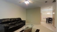 This single-story space presents a straightforward and functional living area finished in warm, neutral tones throughout. The living room is carpeted in a light beige, providing a soft underfoot feel that complements the understated wall color. A ceiling fan with an integrated light fixture is centrally mounted, offering both air circulation and ambient lighting to the room. The space is open and uncluttered, with clean wall lines that create a calm, minimalist atmosphere. A hallway visible in the background leads to additional areas of the home, where a window allows natural light to filter in, brightening the connecting space. The layout feels practical and manageable, well-suited for everyday living. Overall, the space conveys a quiet, no-frills character defined by its neutral palette, comfortable proportions, and simple finishes that provide a solid foundation for personalization.