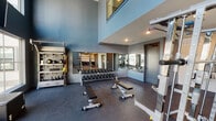 This fitness center is a thoughtfully designed workout facility that combines functionality with sleek, contemporary aesthetics. The space is defined by its dramatic double-height ceilings and floor-to-ceiling windows that flood the interior with natural light while offering views of the surrounding outdoor environment. The dark rubberized flooring throughout provides both durability and a professional gym atmosphere.

The strength training area is generously appointed, featuring a full dumbbell rack with an extensive range of weights, adjustable workout benches, a power rack, and a cable machine with weight plate storage. A wall-length mirror in this section enhances the sense of space and allows for proper form monitoring during exercises. Accessory storage racks along the wall hold kettlebells, medicine balls, and resistance training tools.

The cardio zone, positioned near the expansive window wall, houses treadmills, a stair climber, and stationary cycling bikes, allowing users to enjoy natural light and outdoor views during their workouts. A dedicated spin bike area with additional rowing equipment offers variety for cardiovascular training.

The facility also includes a multi-use open floor area, a built-in hydration station with a dual water fountain set against a white subway tile accent wall, and convenient elevator access. Recessed lighting throughout ensures consistent illumination across all workout zones.