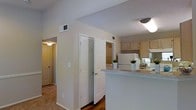 This single-story residence showcases a practical and open layout that connects the main living areas seamlessly. The kitchen is equipped with light wood cabinetry, a white refrigerator, and a peninsula counter finished with a speckled laminate surface, providing additional prep space and a natural dividing point between the kitchen and adjacent areas. The textured ceiling runs throughout the space, adding a subtle visual layer to the interior. Wood-look flooring anchors the kitchen and hallway areas, while plush carpeting defines the living spaces beyond. A six-panel interior door provides access to what appears to be a utility or laundry closet, keeping functional elements neatly tucked away. The hallway connects the main living area to additional rooms, maintaining a smooth flow throughout the floor plan. Neutral wall tones in a warm beige palette contribute to a bright and cohesive atmosphere. Overhead lighting fixtures are present in multiple areas, ensuring consistent illumination across the space. Ventilation is provided via ceiling-mounted HVAC registers. The overall design prioritizes functionality and ease of movement, making it a well-organized and livable single-floor space.