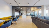 This contemporary single-floor amenity space is thoughtfully designed to accommodate a range of social and functional uses within one expansive, open-plan layout. Large-format tile flooring flows throughout, unifying the distinct zones while contributing to a clean, modern aesthetic.

At the heart of the space sits a substantial kitchen island with a white quartz countertop and integrated seating for multiple guests. The adjacent kitchen wall features rich dark wood cabinetry with gold hardware, a built-in microwave, stainless steel refrigerator, and a sleek white backsplash, providing a well-appointed space for catering and entertaining.

The main lounge area is anchored by a generous sectional sofa in charcoal fabric, positioned beneath a cluster of sculptural woven globe pendant lights. A wall-mounted television and curated botanical artwork complete this relaxed gathering zone. Nearby, a casual dining area offers round tables paired with modern blue accent chairs and yellow upholstered seating for a vibrant pop of color.

A more intimate lounge nook features vertical wood slat wall paneling, open shelving styled with books and greenery, additional woven globe pendants, and low-profile leather seating arranged around a round wooden coffee table — creating a warm, retreat-like atmosphere. Floor-to-ceiling windows at the perimeter flood the space with natural light and offer views to the outdoors.