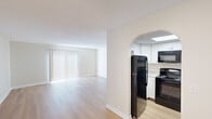 This single-floor residence offers a thoughtfully designed layout that balances open living spaces with private bedroom areas. The living room is a standout feature, characterized by light-toned hardwood-style flooring and a large window dressed with vertical blinds that allow ample natural light to fill the space. A graceful arched doorway connects the living area to the kitchen, creating an open yet defined flow between the two spaces.

The kitchen is efficiently laid out and well-equipped, featuring white cabinetry, granite-look countertops, and a full suite of black appliances including a refrigerator, range, over-the-range microwave, and dishwasher. A skylight overhead provides additional brightness, enhancing the clean, functional aesthetic of the space.

The bedrooms are carpeted and finished in neutral tones, offering a comfortable and quiet atmosphere. Each room includes closet storage—one featuring sliding double doors and another with a bifold door—along with windows fitted with vertical blinds for natural light and privacy. The overall design of the unit is clean and understated, with white trim, neutral walls, and consistent finishes throughout that create a cohesive and move-in ready environment.