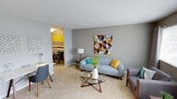 This well-appointed single-floor residence showcases a cohesive and inviting interior design with a mid-century modern aesthetic. The living room serves as the heart of the space, featuring soft, neutral gray walls that complement the plush carpeted flooring. A sleek light-blue sofa paired with colorful accent pillows in teal and yellow anchors the seating area, while a distinctive geometric star-shaped glass coffee table adds a sculptural focal point. A bold geometric wall art piece in warm earth tones adds visual depth and character to the main wall. Thoughtful lighting, including a classic table lamp, ensures the space is warm and welcoming. An open passageway connects the living room to a vibrant dining area, which is distinguished by its striking yellow accent wall that creates a warm, energetic contrast to the otherwise neutral palette. The dining space is furnished with a dark-toned table and chairs, illuminated by overhead lighting that enhances the room's intimate atmosphere. The textured ceiling throughout adds a subtle layer of detail to the overall design. Together, the rooms create a harmonious flow that is both functional and visually appealing, ideal for comfortable everyday living and entertaining.