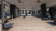 Located at 222 M St SW in Washington, DC, this fitness center offers a spacious, single-floor layout designed with both functionality and modern aesthetics in mind. The space features light wood-tone flooring throughout, creating a clean and inviting atmosphere that contrasts elegantly with the dark structural columns and black-framed glass partitions defining distinct workout zones.

The cardio area is equipped with multiple treadmills and elliptical machines, each featuring individual screens, arranged along floor-to-ceiling mirrored walls that amplify the sense of space. Ceiling fans are strategically positioned overhead to maintain airflow during workouts. Large-scale abstract artwork adorns one wall, adding a striking visual anchor to the open exercise floor.

The strength and functional training zone includes a cable machine, dumbbell rack, adjustable benches, medicine balls, and a TRX suspension training system mounted to a dedicated textured wall panel. A multi-functional cable and pulley rig occupies a dedicated section, offering versatile training options.

The entry area features wood-clad columns, glass-panel doors, and a sanitization station, establishing a polished and professional first impression. Recessed lighting throughout ensures consistent, well-distributed illumination across all areas of the facility.