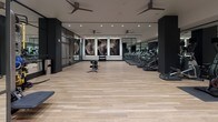 Located at 222 M St SW in Washington, DC, this fitness center offers a spacious, single-floor layout designed with both functionality and modern aesthetics in mind. The space features light wood-tone flooring throughout, creating a clean and inviting atmosphere that contrasts elegantly with the dark structural columns and black-framed glass partitions defining distinct workout zones.

The cardio area is equipped with multiple treadmills and elliptical machines, each featuring individual screens, arranged along floor-to-ceiling mirrored walls that amplify the sense of space. Ceiling fans are strategically positioned overhead to maintain airflow during workouts. Large-scale abstract artwork adorns one wall, adding a striking visual anchor to the open exercise floor.

The strength and functional training zone includes a cable machine, dumbbell rack, adjustable benches, medicine balls, and a TRX suspension training system mounted to a dedicated textured wall panel. A multi-functional cable and pulley rig occupies a dedicated section, offering versatile training options.

The entry area features wood-clad columns, glass-panel doors, and a sanitization station, establishing a polished and professional first impression. Recessed lighting throughout ensures consistent, well-distributed illumination across all areas of the facility.
