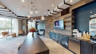 Located at 3202 W Colfax Ave in Denver, Colorado, this single-floor space serves as a polished community or leasing center defined by thoughtful design and high-end material choices throughout. The reception area commands immediate attention with two substantial counters—one featuring dramatic dark, veined stone and another clad in warm, speckled granite—anchoring the room on opposite sides. A long, dark wood central island sits beneath a striking cluster of glass teardrop pendant lights, while an exposed geometric ceiling framework of dark steel and wood beams adds architectural character overhead. Reclaimed wood accent walls introduce a warm, rustic contrast against the contemporary palette of soft grays and blues.

The space transitions seamlessly into a lounge and business center area, where a floor-to-ceiling tiled fireplace wall serves as a bold focal point. Deep navy walls, built-in shelving, and mid-century modern blue upholstered chairs create an inviting, curated atmosphere. Large windows along the perimeter flood the interior with natural light and offer views of the surrounding outdoor area. Neutral tile-patterned carpeting flows throughout, unifying each zone. The overall ambiance balances urban sophistication with approachable warmth, making the space both functional and visually compelling.