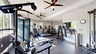 Located at 101 Cambridge Ln in Newtown, PA, this fitness center offers a well-equipped, single-floor exercise space designed to support a comprehensive workout experience. The room features a clean, modern aesthetic with light gray walls accented by bold dark panels, durable rubber flooring, and a tray ceiling with ceiling fans that promote air circulation throughout the space.

Natural light pours in through multiple large windows, providing views of surrounding greenery and creating an energizing atmosphere. The cardio zone is well-stocked with treadmills, elliptical machines, a stair climber, and a stationary bike, all arranged to maximize use of the floor space. A dedicated strength training area houses a multi-station cable machine and a weight bench, ideal for resistance and functional training.

The free weights section features two full dumbbell racks with a wide range of weights, accompanied by a stability ball for accessory exercises. Wall-mounted mirrors in the strength area enhance the sense of space and allow for form checks during workouts. Recessed lighting supplements the natural light, ensuring visibility throughout all hours. Fitness center rules are posted at the entrance, and the space is accessible via a clearly marked exit door. This facility delivers a focused, functional environment well-suited for daily fitness routines.