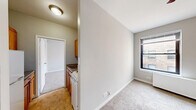 This efficiently designed two-bedroom, one-bathroom unit in Chicago, IL offers a thoughtful layout that maximizes every square foot of its compact footprint. The living area features neutral-toned carpet, a ceiling fan, and a front entry door, creating a welcoming and functional common space that flows directly into the galley-style kitchen. The kitchen is outfitted with warm oak cabinetry, a double-basin stainless steel sink, a coil-burner electric range, and tile flooring, providing all the essentials in a well-organized arrangement. The primary bedroom offers a generous closet with full-width louvered bifold doors, a ceiling fan, and soft carpeting underfoot. The secondary bedroom includes a radiator unit beneath the window, carpet flooring, and direct access to the full bathroom. Each bedroom receives natural light through individual windows with views of the surrounding Chicago streetscape. The bathroom is clean and functional, featuring a full soaking tub with a showerhead, a single vanity with sink, and a mirrored medicine cabinet. Neutral gray and beige tones are carried throughout, lending the space a cohesive and understated aesthetic well-suited for urban living.