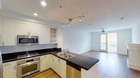 This well-appointed single-floor residence offers a clean, modern aesthetic defined by a cohesive neutral palette and quality finishes throughout. The open-concept layout connects the kitchen directly to the living area, creating a spacious and functional flow. The kitchen is a standout feature, equipped with a full suite of stainless steel appliances—including a side-by-side refrigerator with ice maker, built-in microwave, gas range, and dishwasher—complemented by crisp white cabinetry, glass-front upper cabinet accents, and striking dark granite countertops that extend across the peninsula island. Track lighting overhead adds a contemporary touch. The living area is bathed in natural light from a set of glass doors that open to a private balcony, and is finished with warm hardwood flooring and a ceiling fan. The bathroom maintains the same design language with white cabinetry, dark granite countertops, tile flooring, and a full shower-tub combination. Additional rooms feature the same light hardwood floors and bright white walls, offering versatile, move-in-ready spaces. The overall ambiance is clean, bright, and modern, with a consistent design language that ties the entire residence together seamlessly.