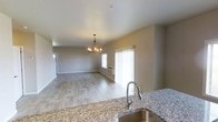 This two-story space offers a thoughtfully designed open-concept main level where the kitchen and living area flow seamlessly together. The kitchen is appointed with granite countertops featuring a speckled finish and a stainless steel sink with a modern pull-down faucet, providing both functionality and style. The countertop extends naturally into the living space, reinforcing the open layout.

The living area is finished with light wood-tone laminate flooring that extends throughout, lending a clean and cohesive aesthetic. Neutral gray walls complement the flooring and contribute to a calm, contemporary atmosphere. A chandelier with multiple arms and warm-toned bulbs serves as a focal point overhead, adding a touch of elegance to the shared space.

Natural light enters through large windows dressed with vertical blinds, brightening the interior. A wall-mounted HVAC unit provides climate control. Smooth white ceilings complete the clean, uncluttered look of the main level. The second floor offers additional living space, expanding the overall footprint of this well-appointed home.