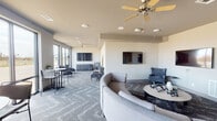 This single-floor communal lounge is thoughtfully designed to accommodate a variety of social settings, offering a versatile and welcoming atmosphere throughout. The space is organized into distinct functional zones, each furnished to encourage both casual relaxation and group interaction.

The primary seating area anchors the room with a generous curved sectional sofa in a neutral gray tone, paired with a round coffee table and complementary accent chairs in blue upholstery. Multiple wall-mounted flat-screen televisions are positioned throughout the space, ensuring clear sightlines from every seating arrangement. A separate nook near the large floor-to-ceiling windows provides a quieter retreat, furnished with a pair of high-back lounge chairs, a side table, and a dedicated wall-mounted TV, all bathed in abundant natural light with views of the surrounding exterior landscape.

Toward the opposite end of the room, high-top bistro tables and chairs offer a more upright setting suited for casual dining or socializing. Interior pass-through windows along one wall hint at an adjacent space, likely a kitchen or service area. Patterned chevron-style carpet in cool gray tones flows throughout, tying the zones together cohesively. Ceiling fans with warm-toned wood blades and recessed lighting complete the clean, modern aesthetic.
