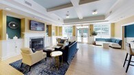 This single-floor clubhouse is thoughtfully designed to serve as a welcoming communal gathering space. The main lounge area is the centerpiece of the facility, featuring a classic white-mantled fireplace, a wall-mounted flat-screen television, and a coffered ceiling that adds architectural character. Warm golden-yellow walls, wainscoting, and light hardwood flooring create an inviting ambiance, while large multi-pane windows and glass double doors flood the space with natural light and offer views of the surrounding greenery and pool area. Stylish blue upholstered sofas and accent chairs are arranged for comfortable socializing.

The fully equipped kitchen includes an abundance of white cabinetry, dark granite countertops, stainless steel appliances — including a full-size refrigerator, dishwasher, and microwave — as well as a sink, all set on durable tile flooring. An ADA-accessible lowered counter section is also incorporated into the design.

A dedicated business center offers workstations with desktop computers and a printer, set against bold teal accent walls with white wainscoting and patterned carpet. Adjacent to it is a private meeting room featuring the same teal and white color palette, a large window, decorative artwork, a dining table, and a pendant chandelier — ideal for small gatherings or focused work sessions.