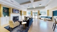 This single-floor clubhouse is thoughtfully designed to serve as a welcoming communal gathering space. The main lounge area is the centerpiece of the facility, featuring a classic white-mantled fireplace, a wall-mounted flat-screen television, and a coffered ceiling that adds architectural character. Warm golden-yellow walls, wainscoting, and light hardwood flooring create an inviting ambiance, while large multi-pane windows and glass double doors flood the space with natural light and offer views of the surrounding greenery and pool area. Stylish blue upholstered sofas and accent chairs are arranged for comfortable socializing.

The fully equipped kitchen includes an abundance of white cabinetry, dark granite countertops, stainless steel appliances — including a full-size refrigerator, dishwasher, and microwave — as well as a sink, all set on durable tile flooring. An ADA-accessible lowered counter section is also incorporated into the design.

A dedicated business center offers workstations with desktop computers and a printer, set against bold teal accent walls with white wainscoting and patterned carpet. Adjacent to it is a private meeting room featuring the same teal and white color palette, a large window, decorative artwork, a dining table, and a pendant chandelier — ideal for small gatherings or focused work sessions.