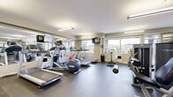 Located at 235 Willis Cres in Saskatoon, Saskatchewan, this fitness facility offers a well-appointed exercise room designed to support a complete workout regimen. The space is laid out on a single floor and provides generous room to accommodate a wide variety of equipment without feeling crowded.

The cardio zone features multiple treadmills, elliptical trainers, stationary bikes, and additional cardio machines, all positioned to take advantage of the natural light streaming in through large windows that frame views of the surrounding neighbourhood. Wall-mounted televisions are conveniently placed for entertainment during workouts.

The strength training area is equally comprehensive, with a full rack of free weights and dumbbells, a multi-station cable machine, weight benches, a leg press, and additional resistance machines. Full-length mirrors line the walls, visually expanding the space while allowing users to monitor their form throughout their routines.

Durable rubber flooring covers the entire exercise area, providing safety and comfort underfoot. Fluorescent lighting ensures the room remains well-lit at all times. The clean, neutral colour palette and organized equipment arrangement contribute to a focused and motivating training environment, making this fitness facility a practical and fully functional amenity.