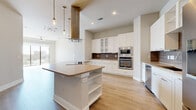 This thoughtfully designed single-floor residence offers a clean, contemporary aesthetic throughout. The heart of the home is an expansive open-concept kitchen, anchored by a large central island with a warm countertop and built-in shelving. A striking brass island range hood serves as a focal point, complemented by white shaker-style cabinetry, glass-front upper cabinets, a textured tile backsplash, and a full suite of stainless steel appliances including a double wall oven, built-in microwave, and French door refrigerator. Light wood-look flooring flows seamlessly through the main living areas, enhancing the bright, airy atmosphere. The living space opens to a private balcony through floor-to-ceiling sliding glass doors, framing impressive city skyline views and filling the room with abundant natural light. Ceiling fans are present throughout for added comfort. The bedrooms feature plush carpeting and generous proportions, with one bedroom offering its own city-facing window and ceiling fan. A spacious walk-in closet provides ample built-in shelving and hanging rods. The bathroom is finished with large-format tile flooring, a dual-sink vanity with white cabinetry, and a glass-enclosed walk-in shower. A dedicated laundry room accommodates a full-size side-by-side washer and dryer with overhead shelving.