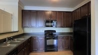 Located at 5806 Ogeechee Rd in Savannah, Georgia, this single-story property offers a comfortable and functional layout designed for everyday living. The kitchen is a standout feature of the space, showcasing rich, dark espresso-toned cabinetry that extends to the ceiling and is accented with sleek hardware. Crown molding adds a refined finishing touch along the upper edge of the cabinetry, lending a sense of polish to the room. The kitchen is equipped with black appliances, including a freestanding electric range, an over-the-range microwave, and a refrigerator, all seamlessly integrated into the cabinetry layout. Dark countertops provide a striking contrast against the lighter wall tones, while warm hardwood-style flooring grounds the space with a welcoming feel. Ample upper and lower cabinet storage ensures generous organization options throughout the kitchen. The overall aesthetic is cohesive and contemporary, with a neutral color palette that creates a clean, inviting atmosphere. The single-floor design promotes ease of movement and accessibility throughout the home, making it a practical and appealing option in the Savannah, GA area.