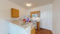 Located at 4510 Tesla Park Dr in Wilmington, NC, this single-floor unit offers a practical and well-organized layout designed for comfortable everyday living. The kitchen is a functional highlight, featuring warm honey-toned cabinetry, white appliances including a coil-burner range, over-the-range microwave, and refrigerator. A peninsula breakfast bar with a light countertop extends into the living area, creating an open, connected feel between the kitchen and main living space. The kitchen is finished with durable hardwood-style flooring that adds warmth and is easy to maintain. The living area is generously proportioned, carpeted in neutral tones, and fitted with a ceiling fan for year-round comfort. A door with a transom window allows natural light to fill the space, adding brightness to the open floor plan. An additional carpeted room with its own ceiling fan serves well as a bedroom or flex space, offering a clean, versatile canvas. The bathroom is neatly appointed with a single-sink vanity featuring light wood cabinetry, a wall mirror with overhead lighting, and a shower/tub combination. Throughout the unit, the bright white walls and consistent neutral palette create a clean, cohesive ambiance.